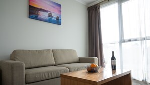 Un salón con un sofá color beige, una mesa de centro de madera con una botella de vino y copas, y un cuadro de un atardecer sobre rocas en la pared.