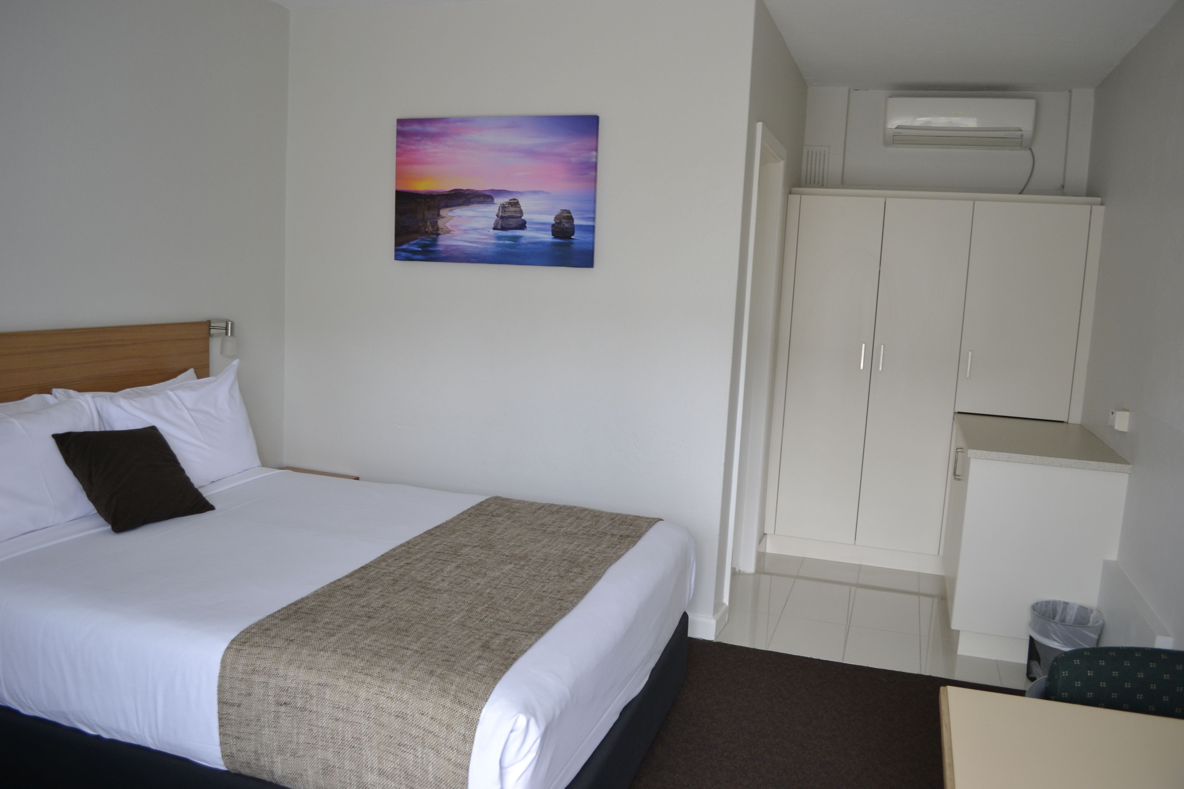 A hotel room with a bed, a painting of a sunset, a wardrobe, and an air conditioning unit.