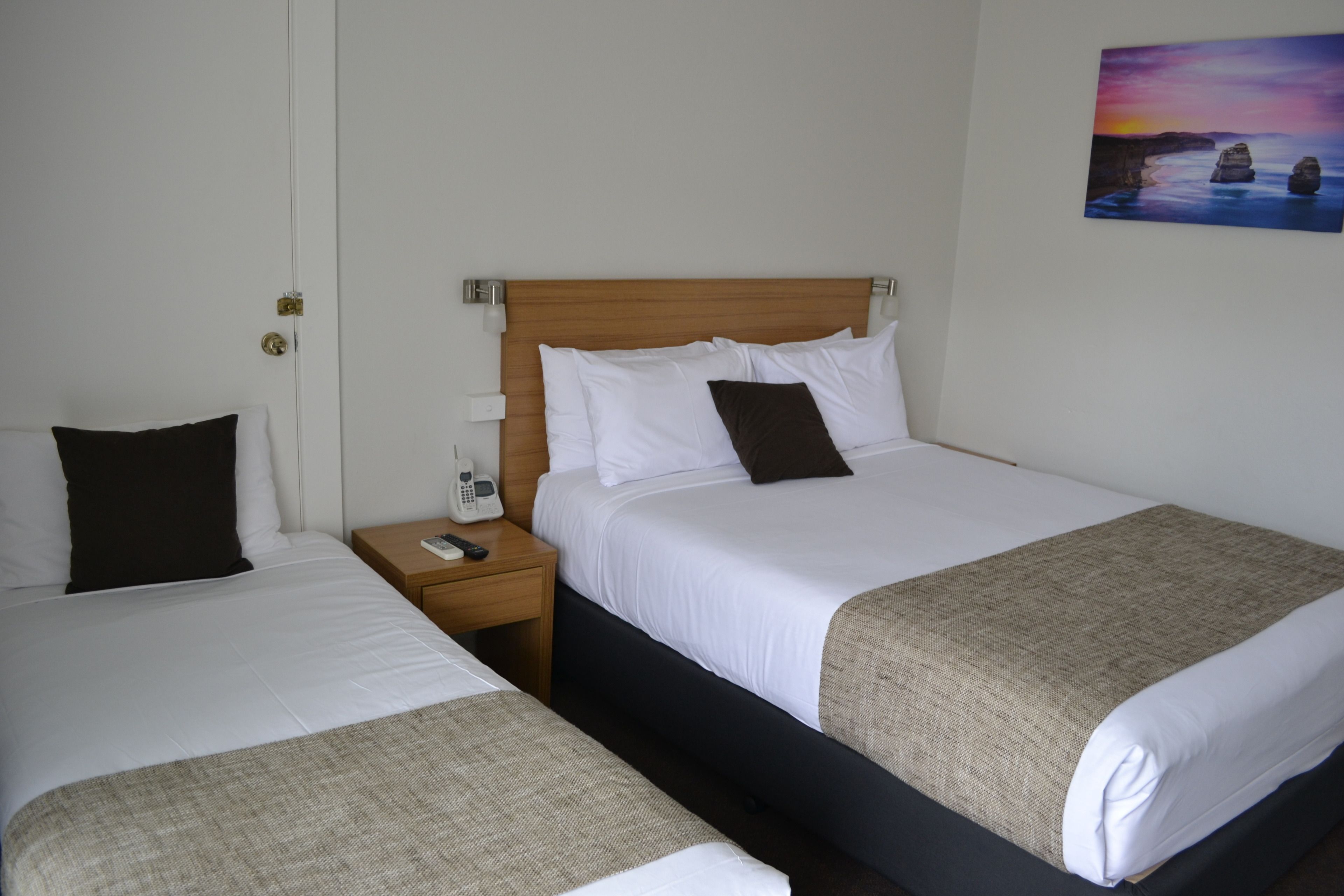 A hotel room with two single beds, a wooden headboard, a nightstand with a phone, and a painting on the wall.