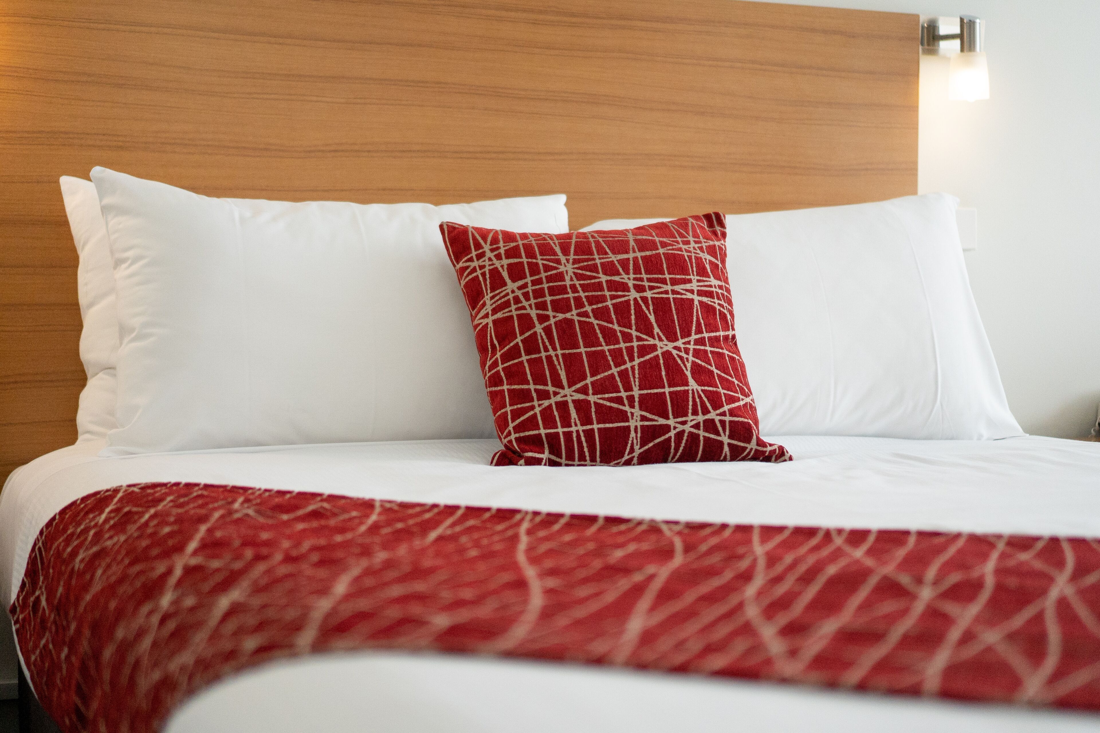 Un lit bien fait, avec une literie blanche et rouge, une tête de lit en bois et un luminaire fixé au mur.
