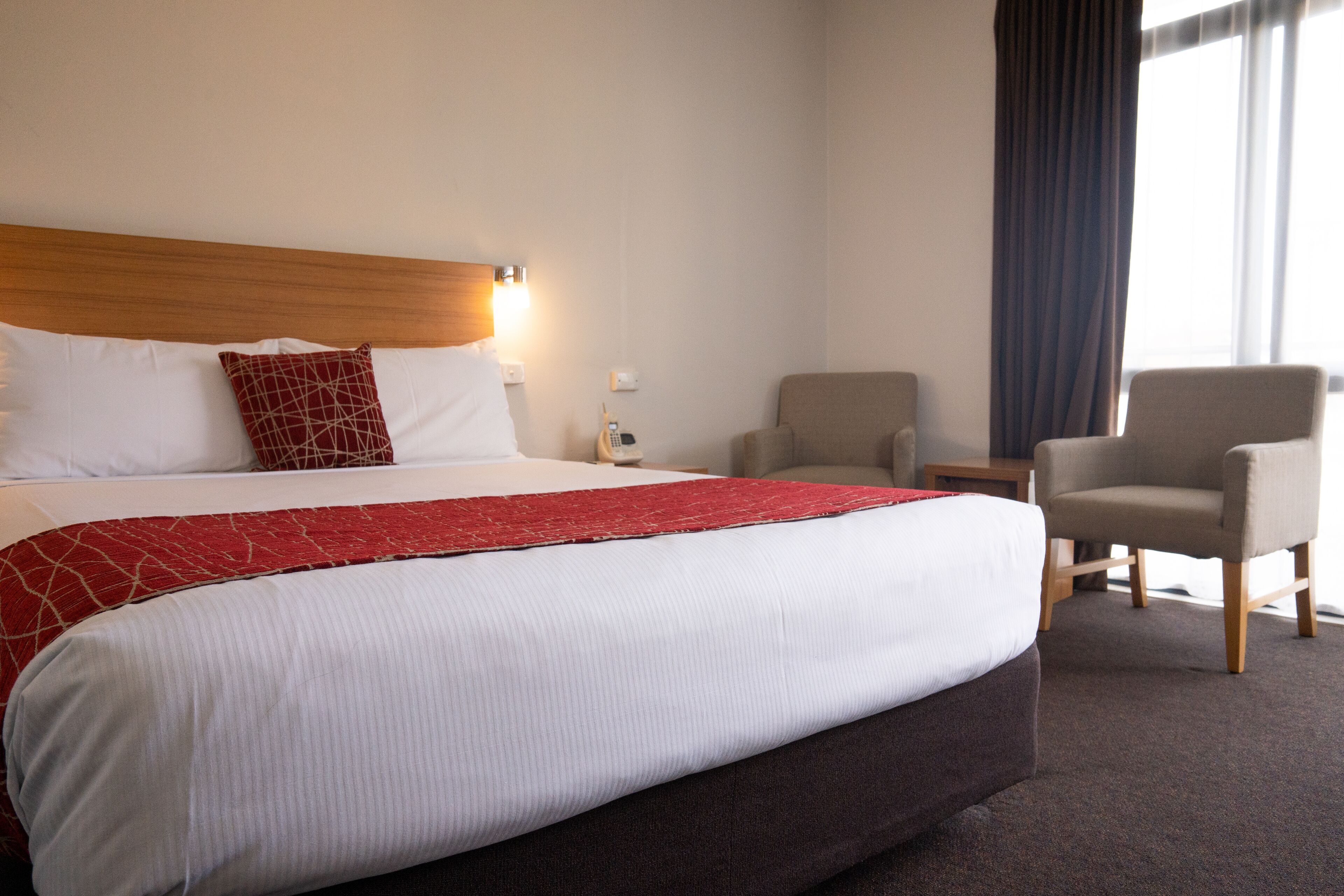 Une chambre d’hôtel avec un grand lit, une tête de lit en bois, une couvre-lit rouge et blanche, un fauteuil gris et une petite table avec une lampe.