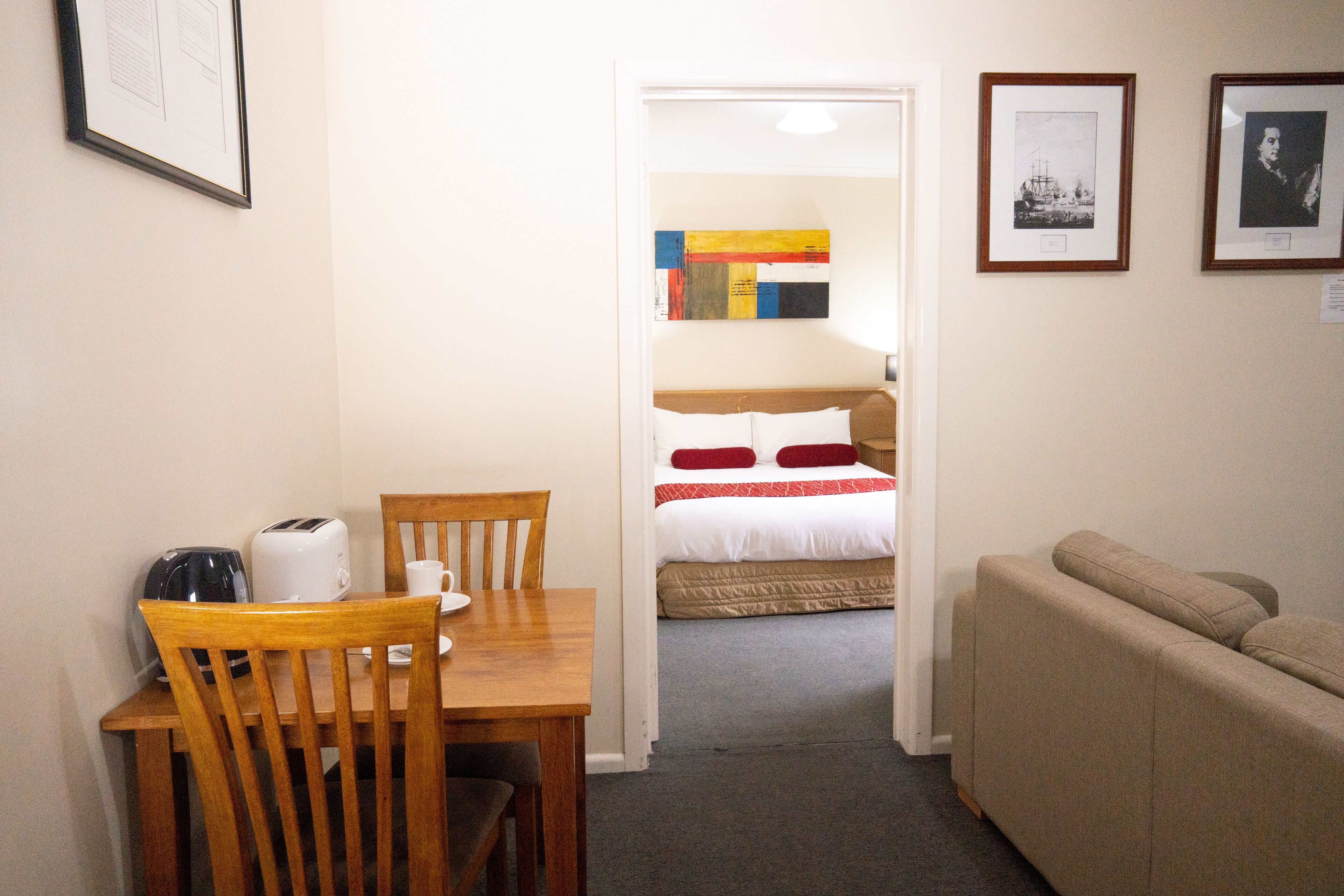 A room with a wooden dining table, chairs, a bed with a red and white bedspread, and a beige sofa.