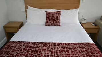 Una cama bien hecha con una colcha de cuadros rojos y blancos, almohadas blancas y un cabecero de madera. Hay dos mesitas de noche, una con una lámpara blanca y la otra con un teléfono blanco y un control remoto.