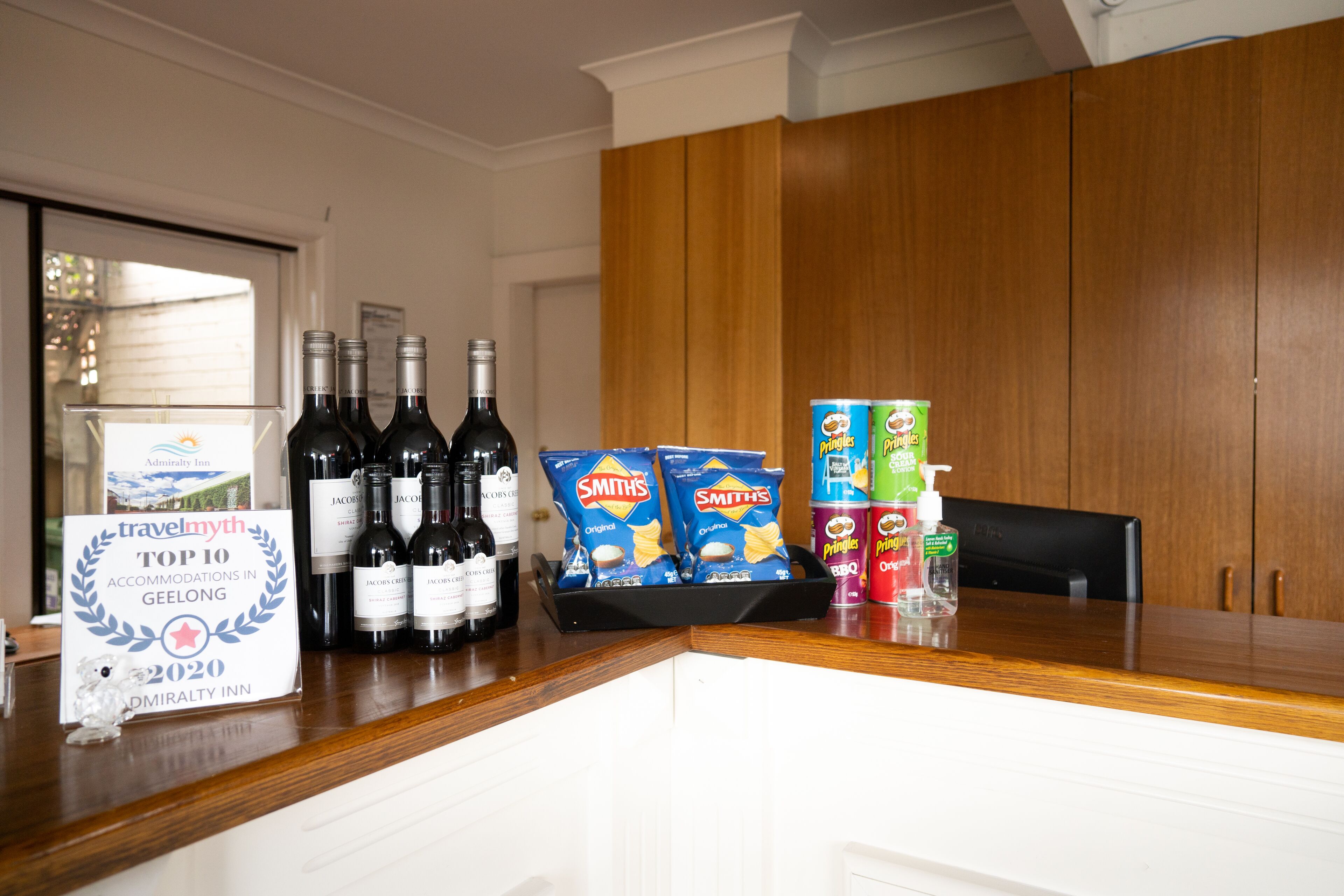A hotel reception area with a wooden counter, bottles of wine, snacks, and a sign listing top accommodations in Geelong for 2020.