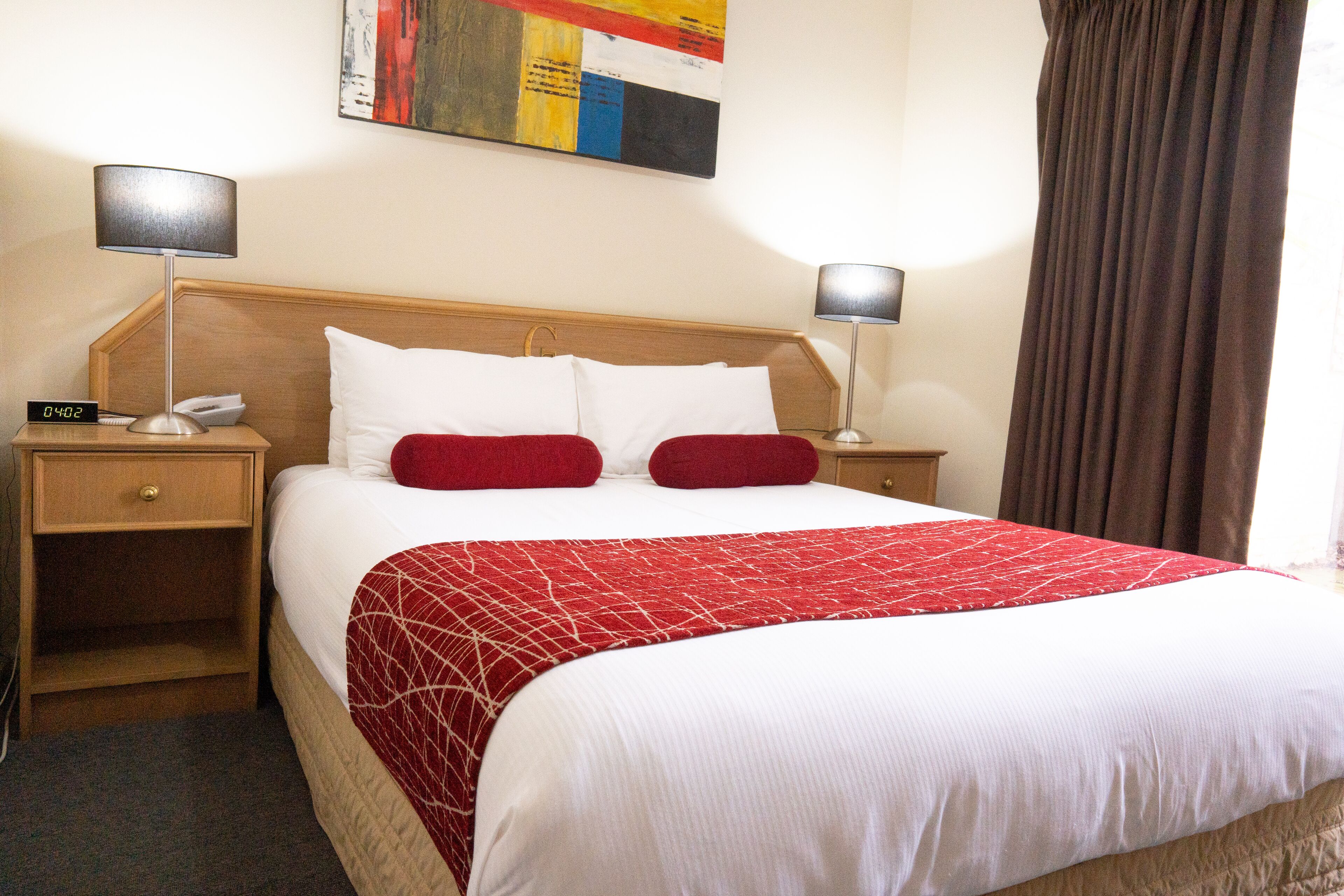 A hotel room with a large bed, two bedside tables, a nightstand, a red and white bedspread, and a painting on the wall.