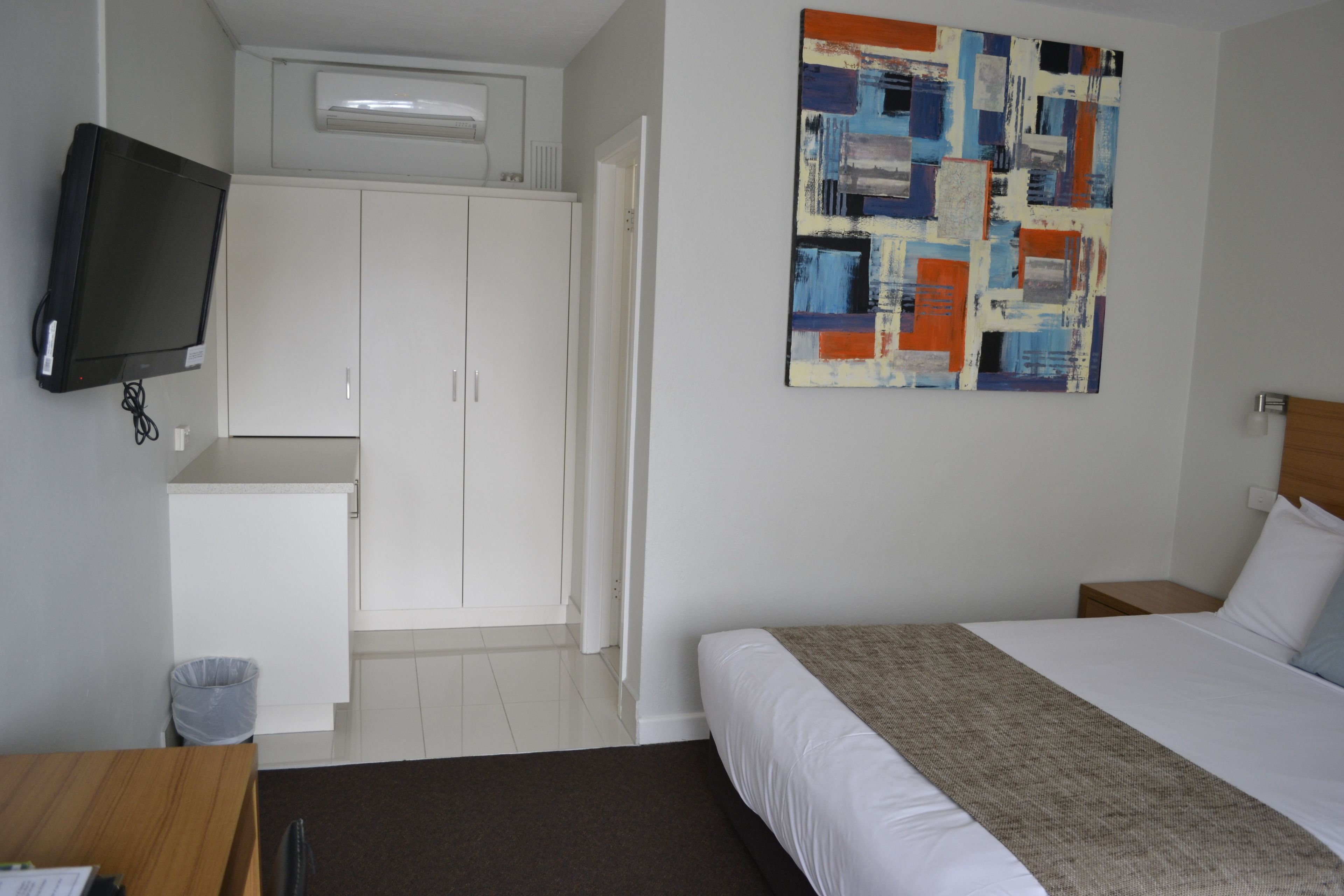 Habitación de hotel con cama, televisor montado en la pared, armario y un cuadro.