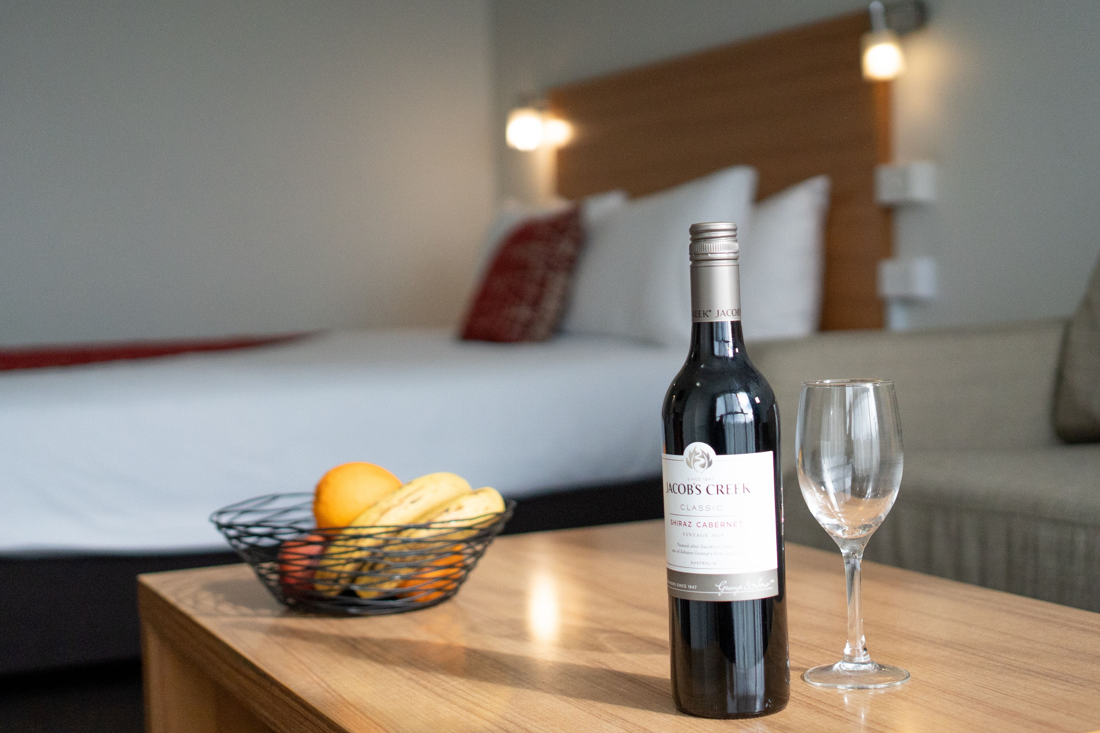 A bottle of wine and a glass on a wooden table, with a basket of fruit and a bed in the background.