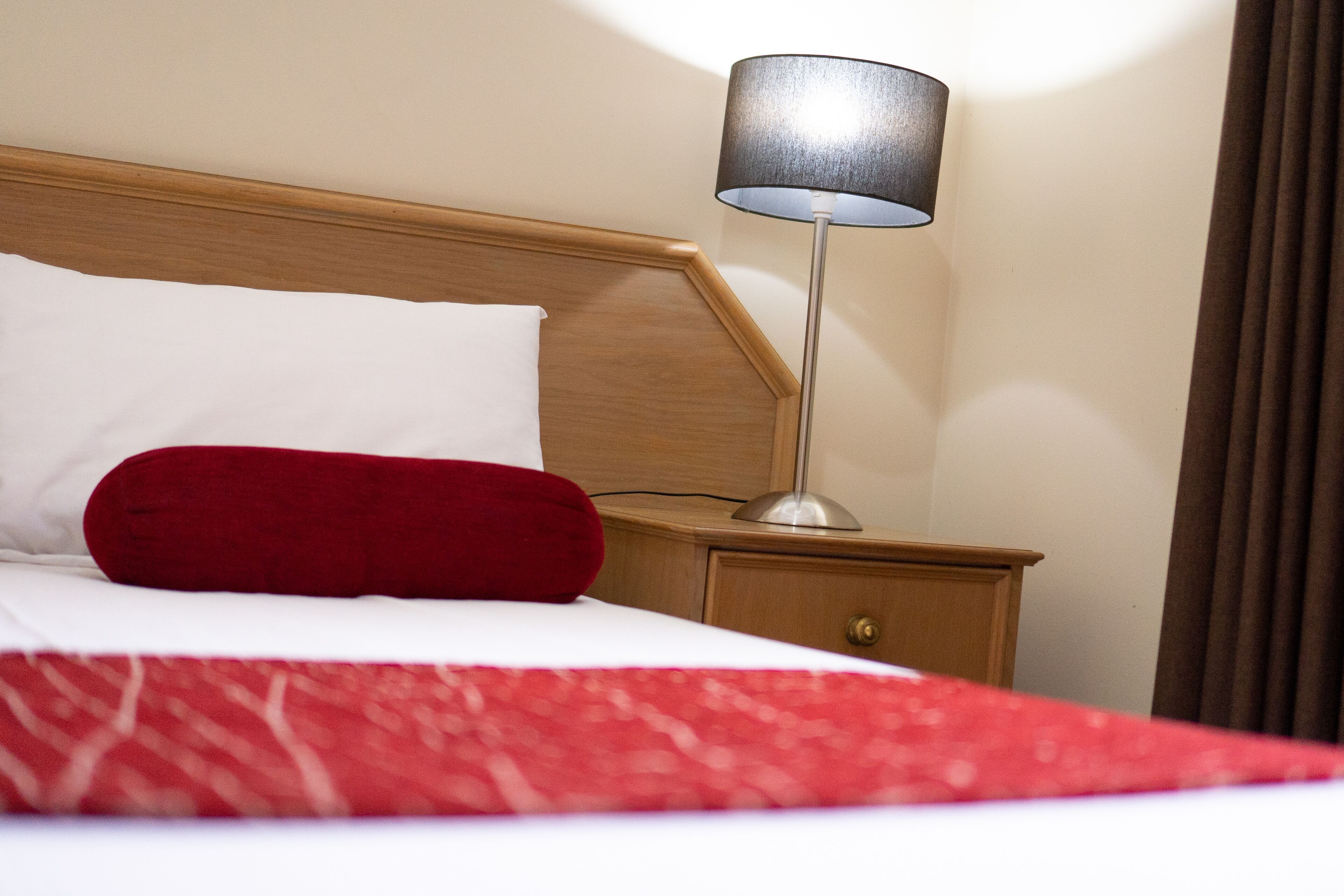 A neatly made bed with a red blanket and pillow, a bedside lamp, and a wooden nightstand.