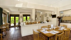 Un restaurante con mesas y sillas de madera, una barra de buffet con exhibiciones de comida, y vistas al exterior con vegetación.