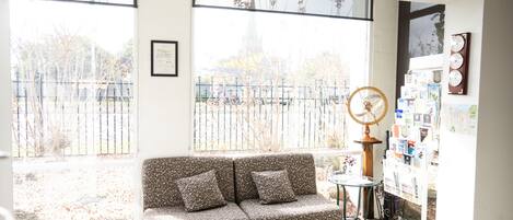 A room with a sofa, a small table, and a wall-mounted fan. There is a window with a view of a fence and trees.