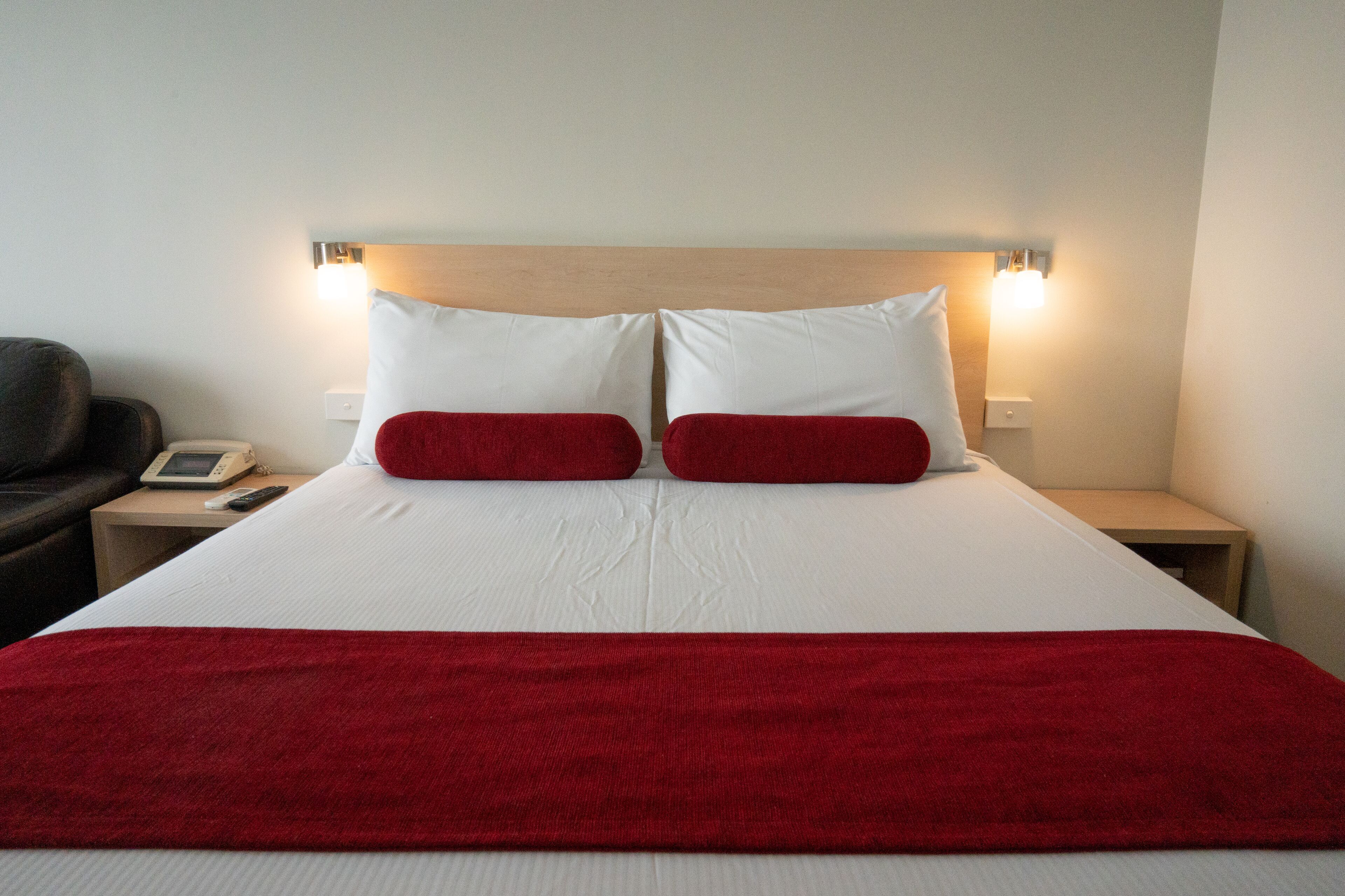 Una cama bien tendida con ropa de cama blanca y roja, dos mesitas de noche con lámparas, y una silla a la izquierda.