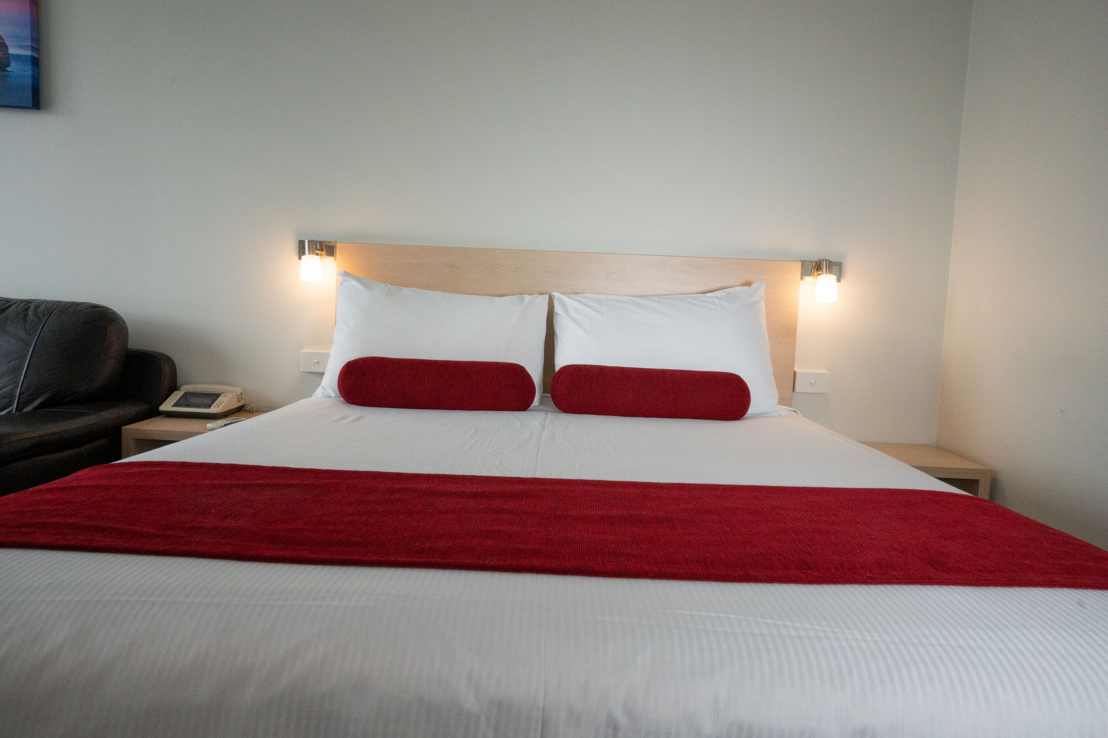 A hotel room with a large bed, two red pillows, a wooden headboard, bedside lamps, a telephone, and a red throw blanket.