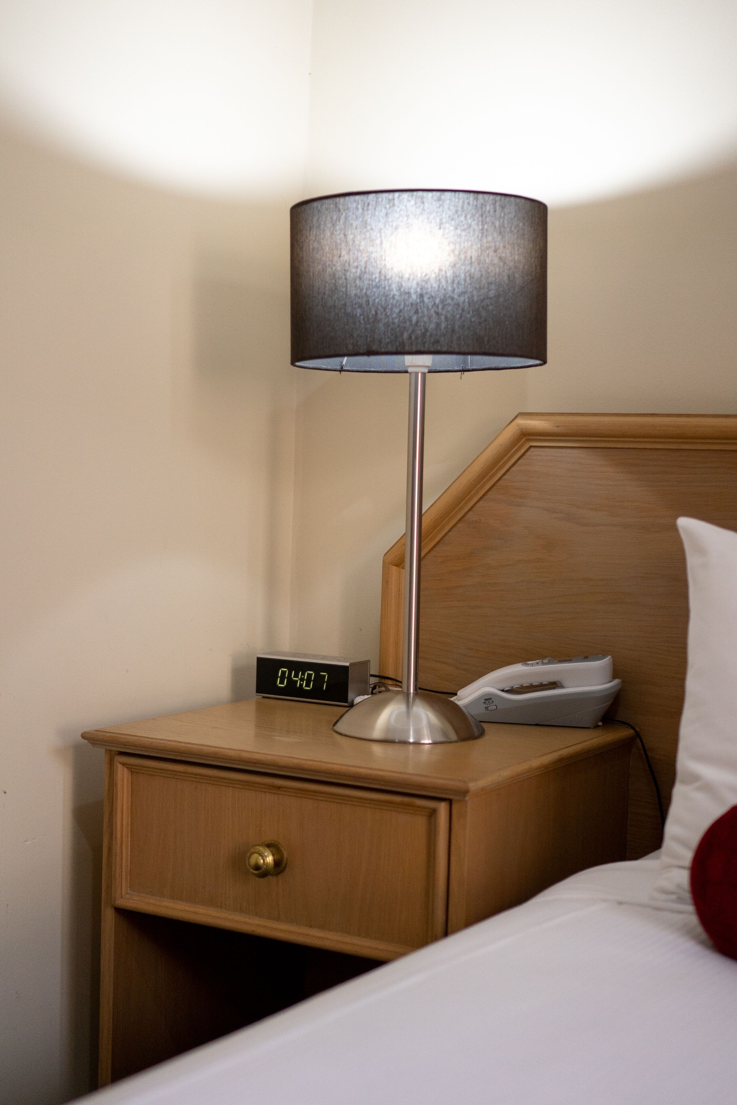 A wooden bedside table with a digital clock displaying 04:07, a lamp, and a telephone.