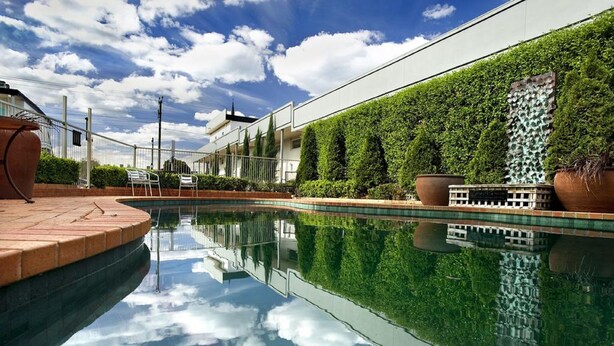 Una piscina con reflejo cristalino, rodeada de un seto cuidado y un edificio moderno de techo plano.
