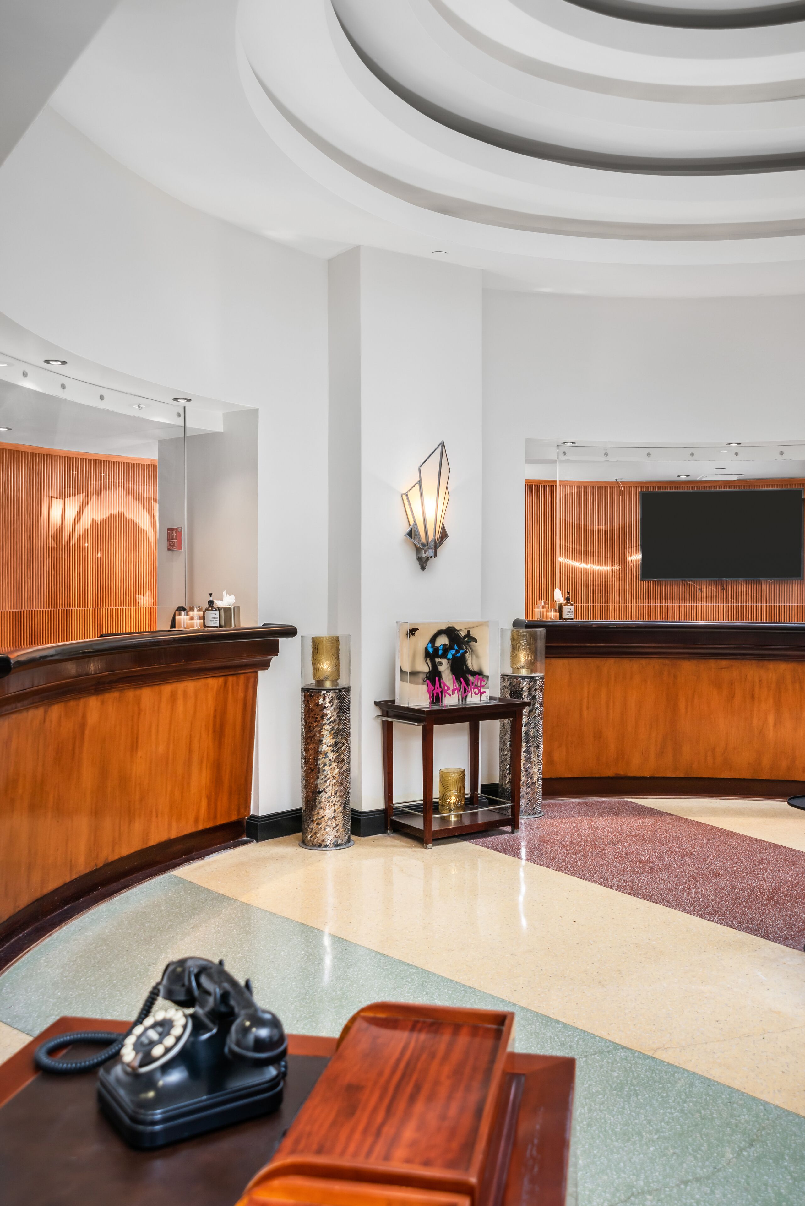 A hotel reception area with a wooden reception desk, a telephone, a small table with decorative items, a wall-mounted TV, and a patterned floor.
