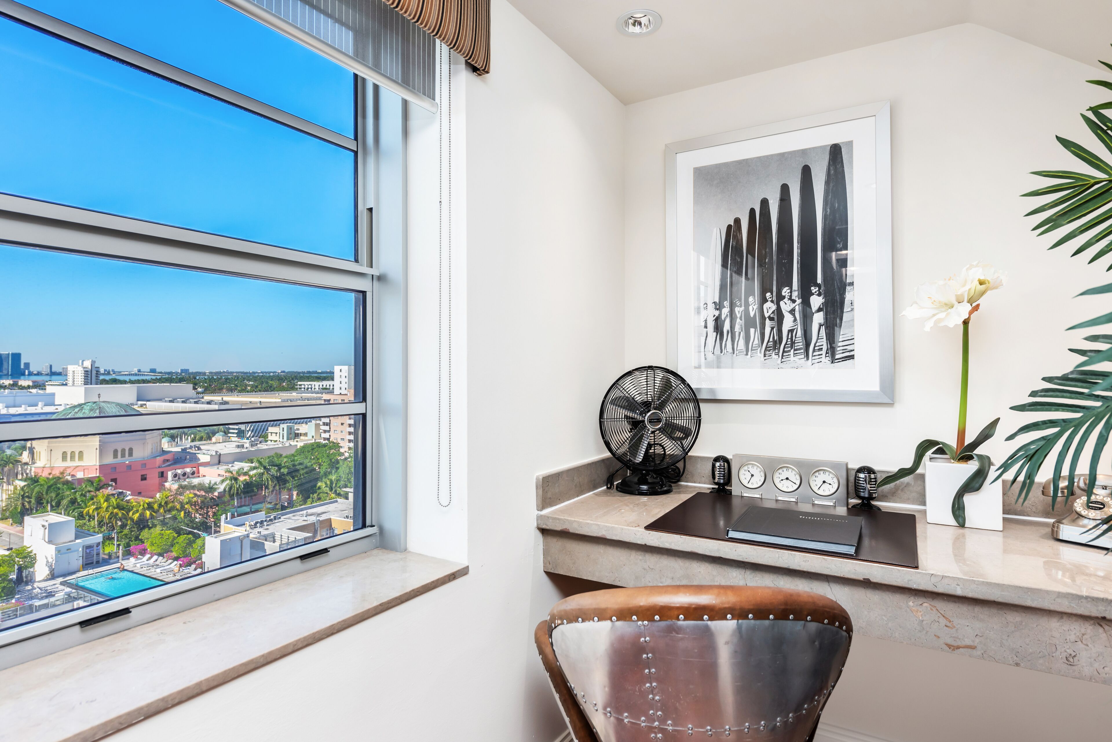 A modern room with a large window, a desk with a fan, a chair, a clock, and a framed artwork.