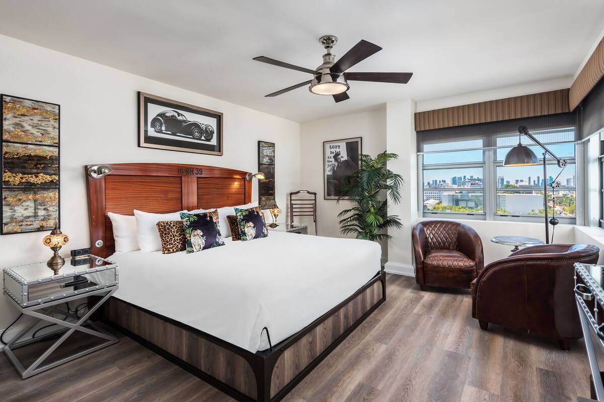 A modern hotel room with a large bed, a ceiling fan, a city view, and a leather armchair.