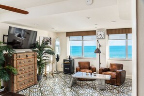 A spacious living room with a large window offering a view of the ocean, a vintage suitcase, a Marshall amplifier, and a leather armchair.