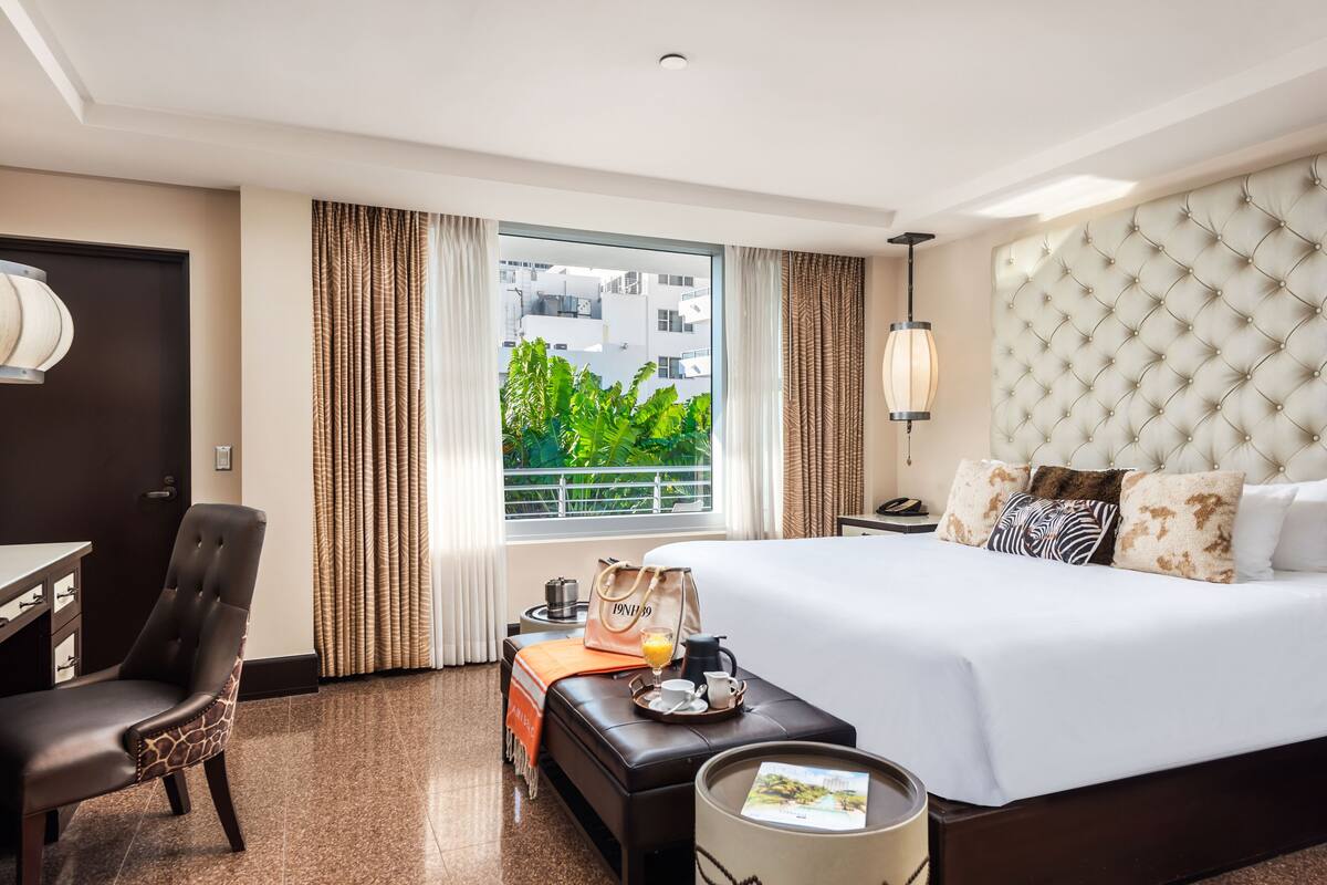 A hotel room with a large bed, a desk, a chair, a window with curtains, and a view of buildings and greenery outside.