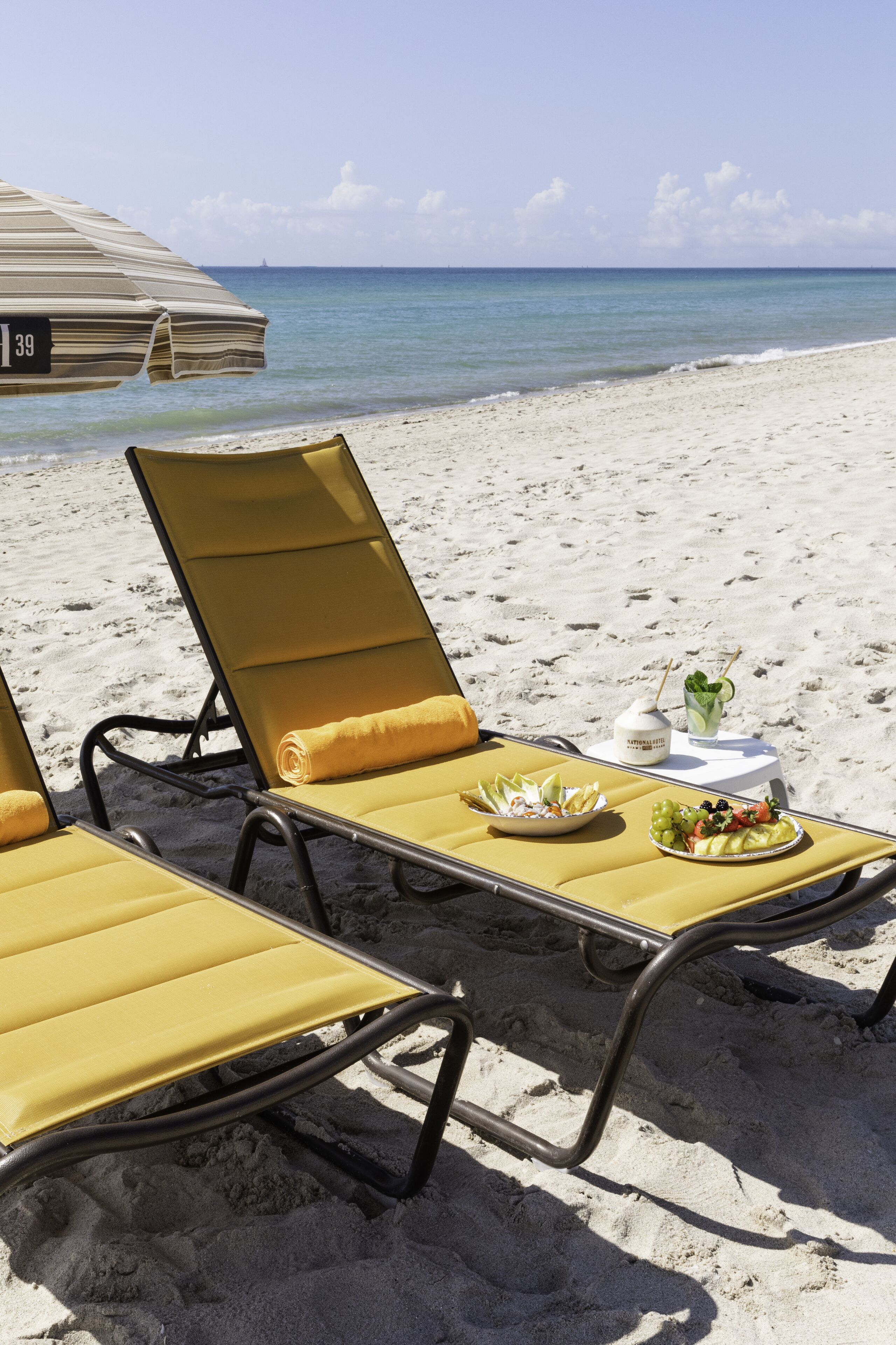 Un área de descanso en la playa con dos sillones amarillos, una mesa con fruteros y bebidas, y una sombrilla de playa a rayas.