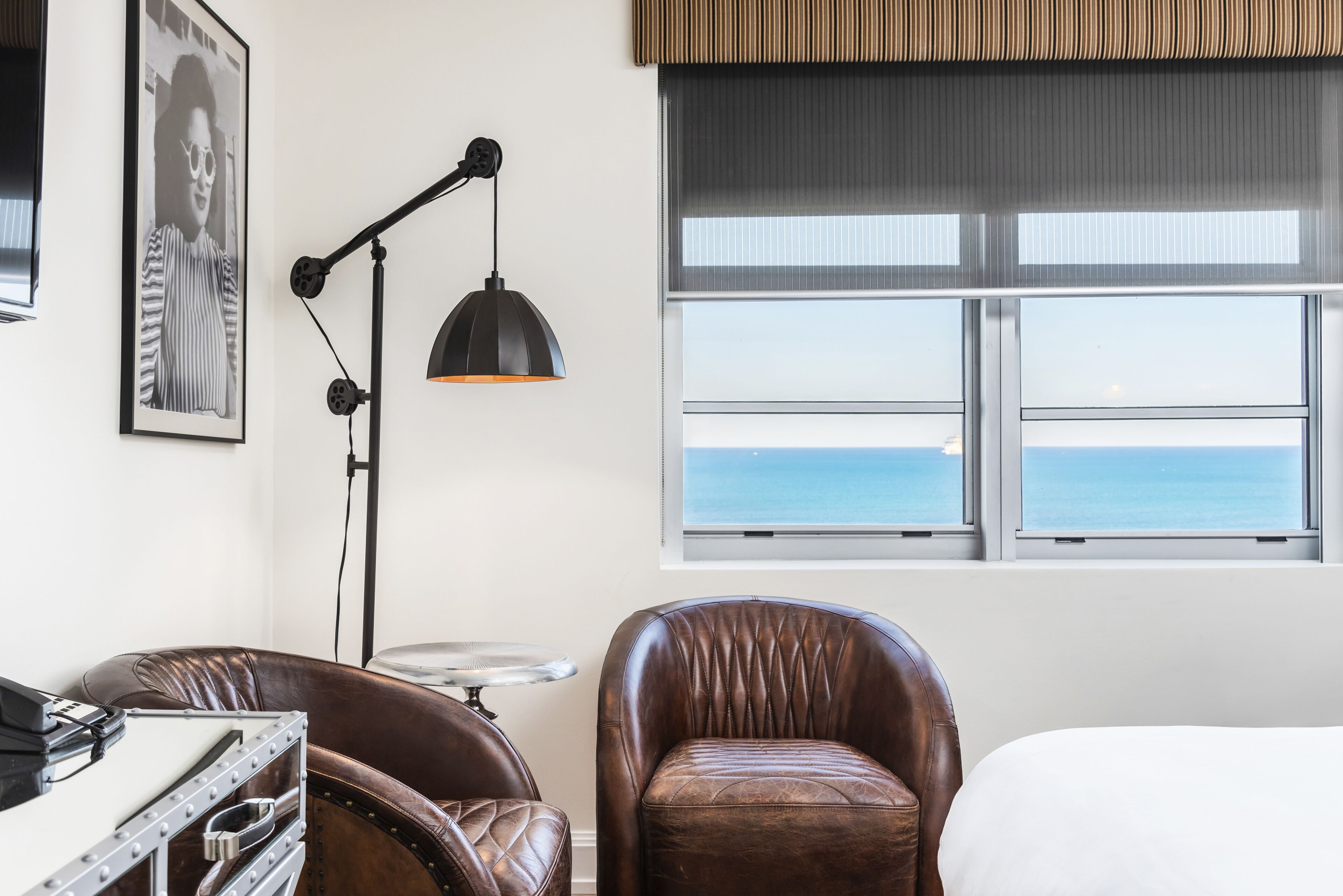 Ein Hotelzimmer mit Meerblick, einem Schreibtisch mit Lampe, einem Ledersessel und einem Bett.
