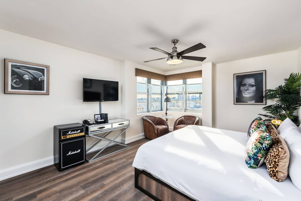 A hotel room with a bed, a desk with a television, a ceiling fan, and a large window with a city view.