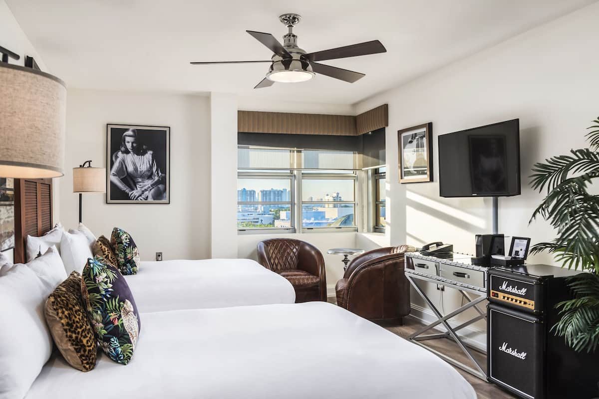 A hotel room with two beds, a ceiling fan, a television, a Marshall amplifier, and a city view.
