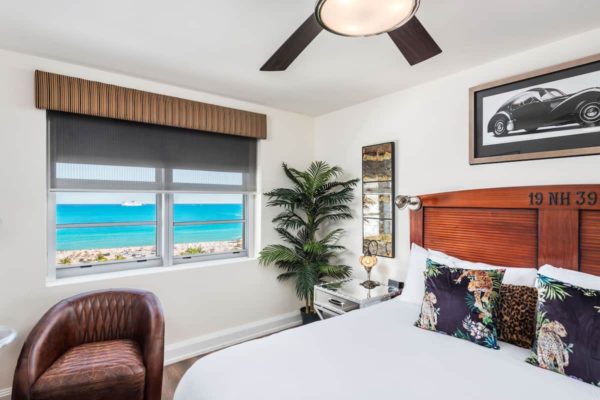 A hotel room with a large window offering a beach view, a ceiling fan, a bed with leopard print pillows, and a framed car picture on the wall.