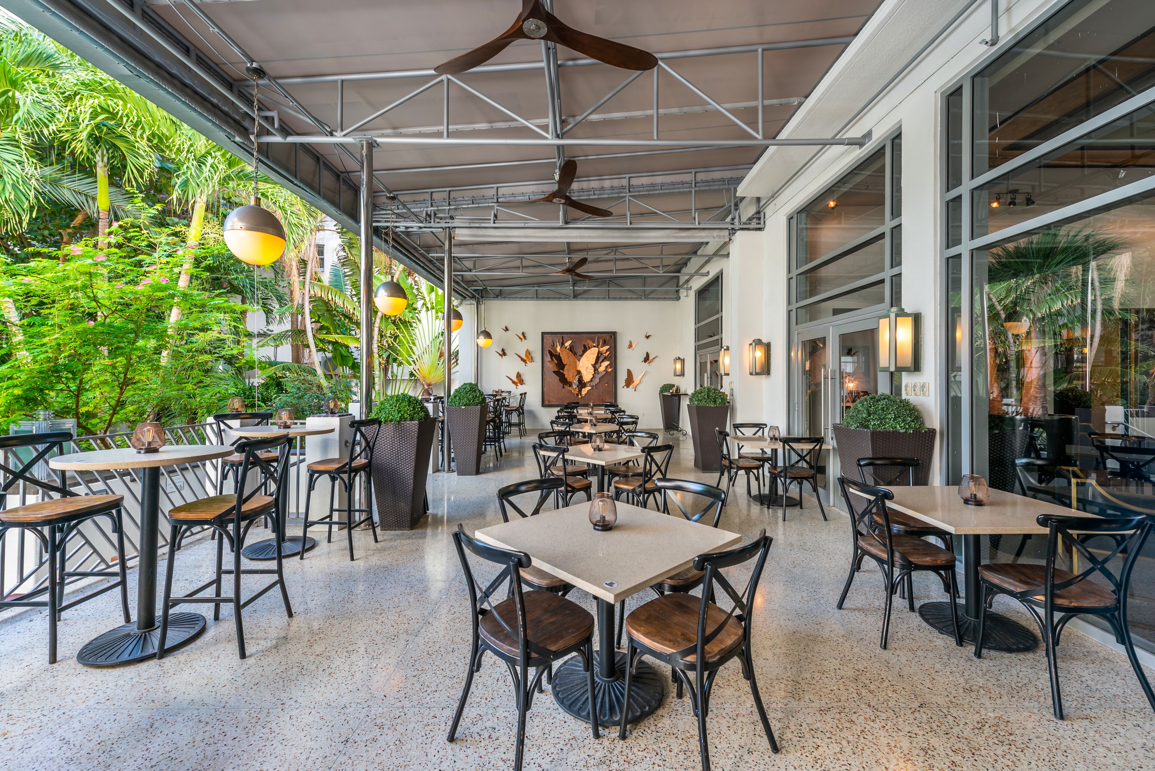 An indoor dining area with multiple tables and chairs, ceiling fans, and large windows overlooking a lush garden.