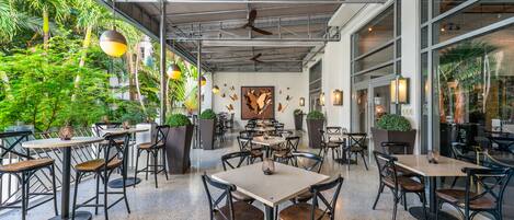 An indoor dining area with multiple tables and chairs, ceiling fans, and large windows overlooking a lush garden.