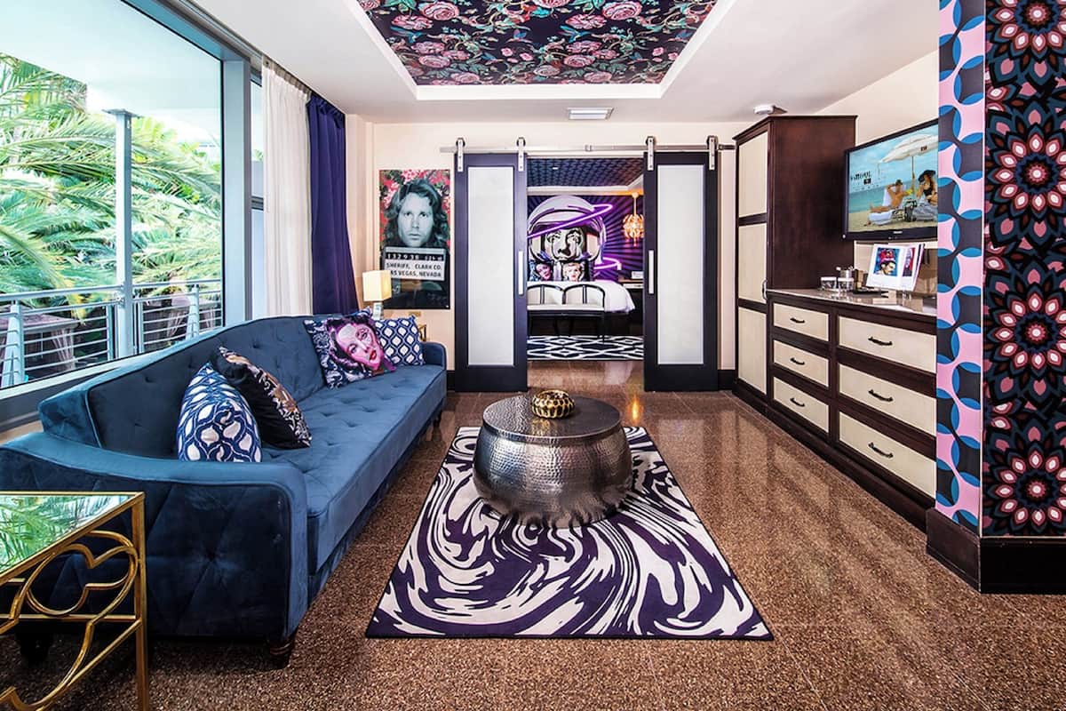 A modern hotel room with a blue sofa, a unique coffee table, a patterned rug, and a view of palm trees.