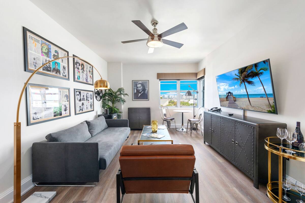 A modern living room with a gray sofa, a brown ottoman, a ceiling fan, and a large TV screen displaying a beach scene.