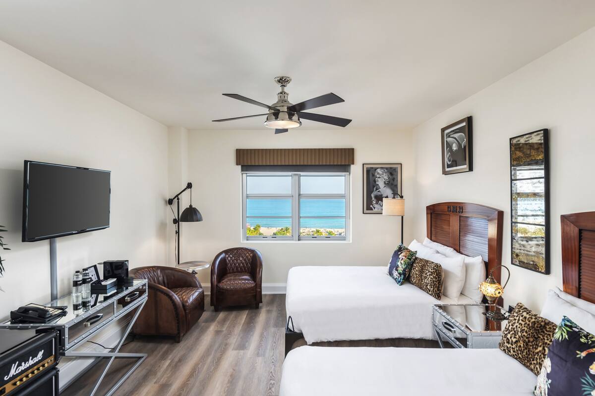 A hotel room with two beds, a TV, a desk with a computer and speakers, a chair, and a window with a view of the beach.