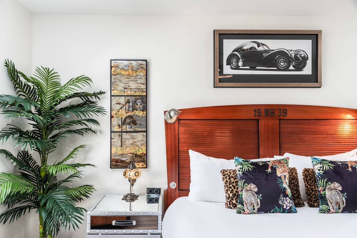 A bedroom with a wooden headboard, a large potted plant, a framed car picture, and a wall art piece.