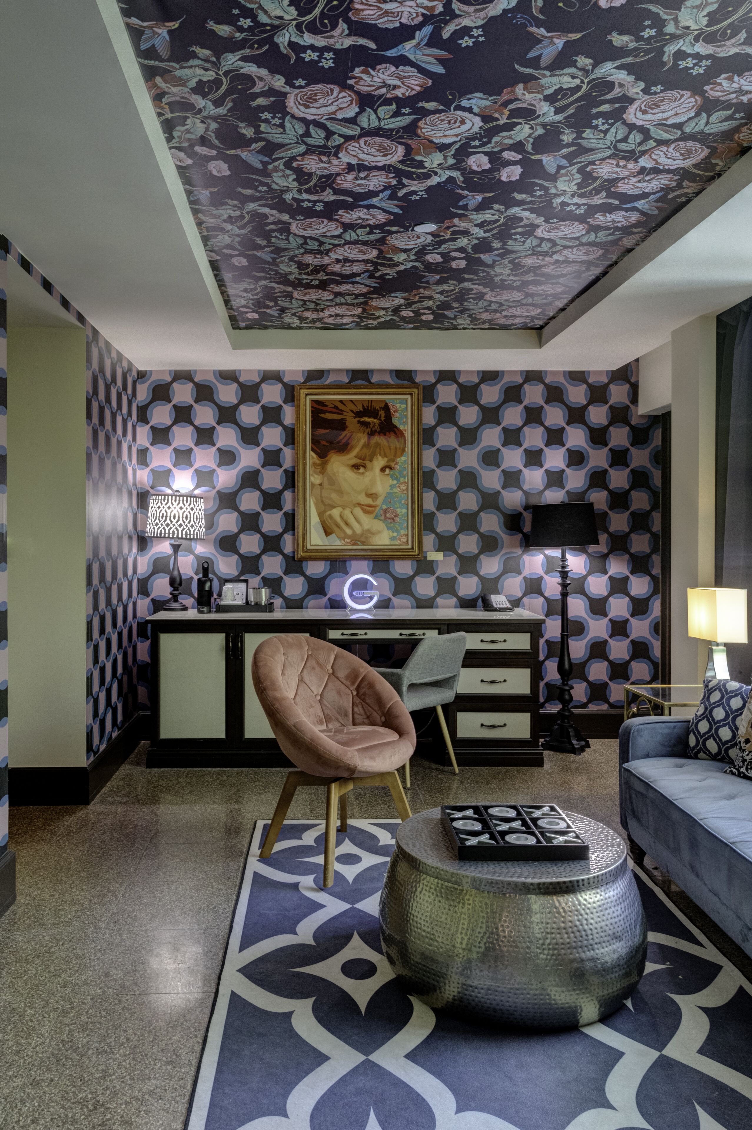 A modern living room with a checkered wall pattern, a floral ceiling, a desk with a lamp, a chair, and a round coffee table.