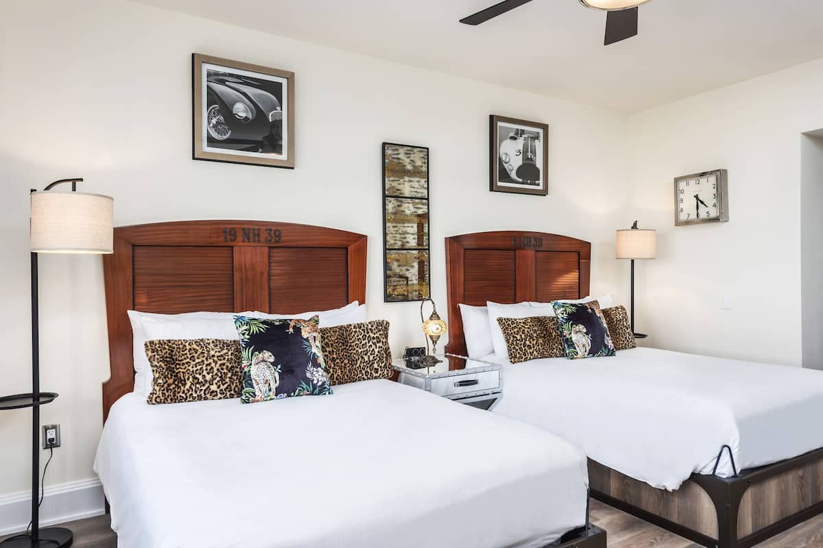 A hotel room with two beds, each with a wooden headboard, white bedding, and leopard print pillows. There are wall-mounted clocks and framed pictures above the beds.