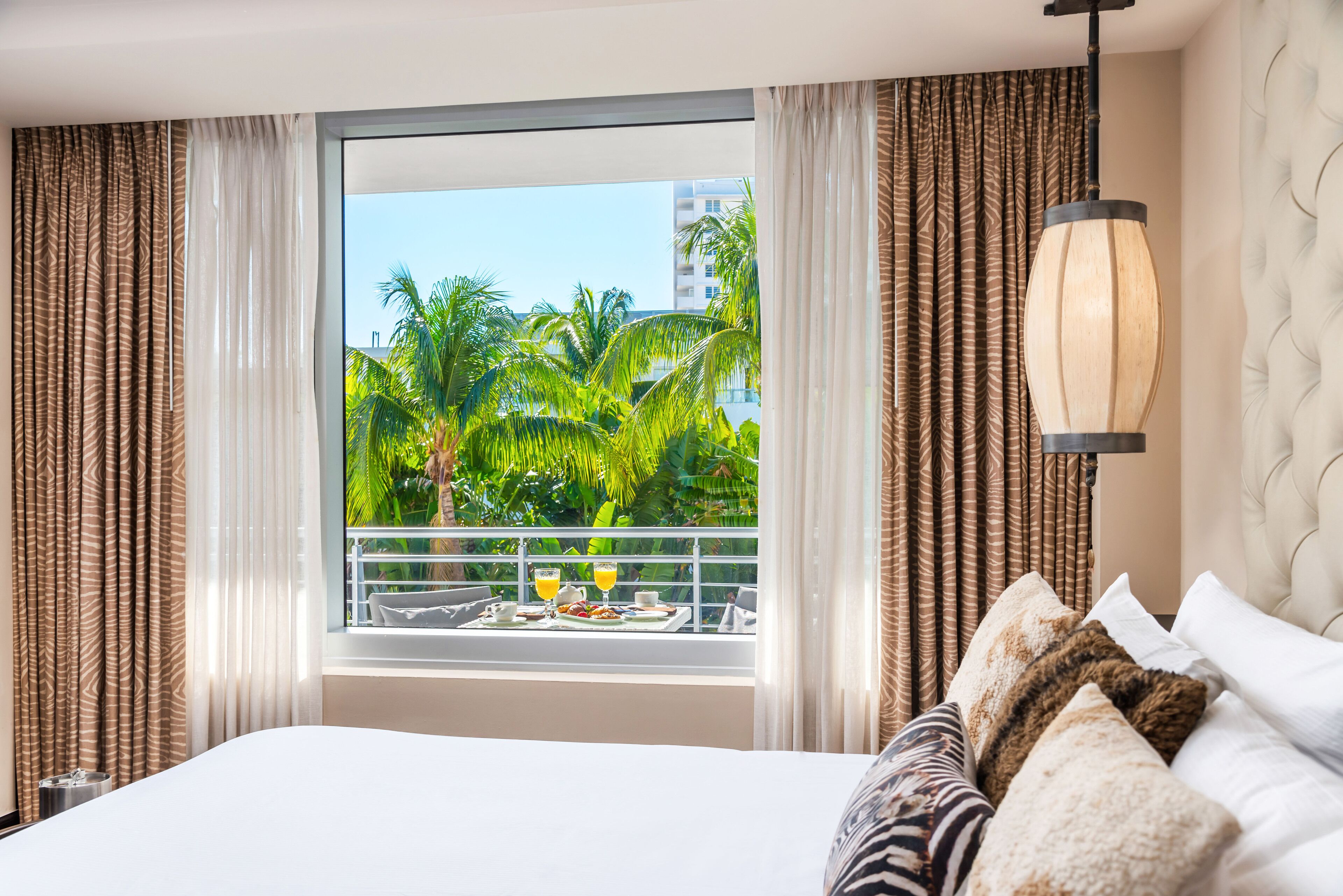 Quarto de hotel com uma cama grande, janela com cortinas, varanda com mesa posta para café da manhã e luminária pendente.