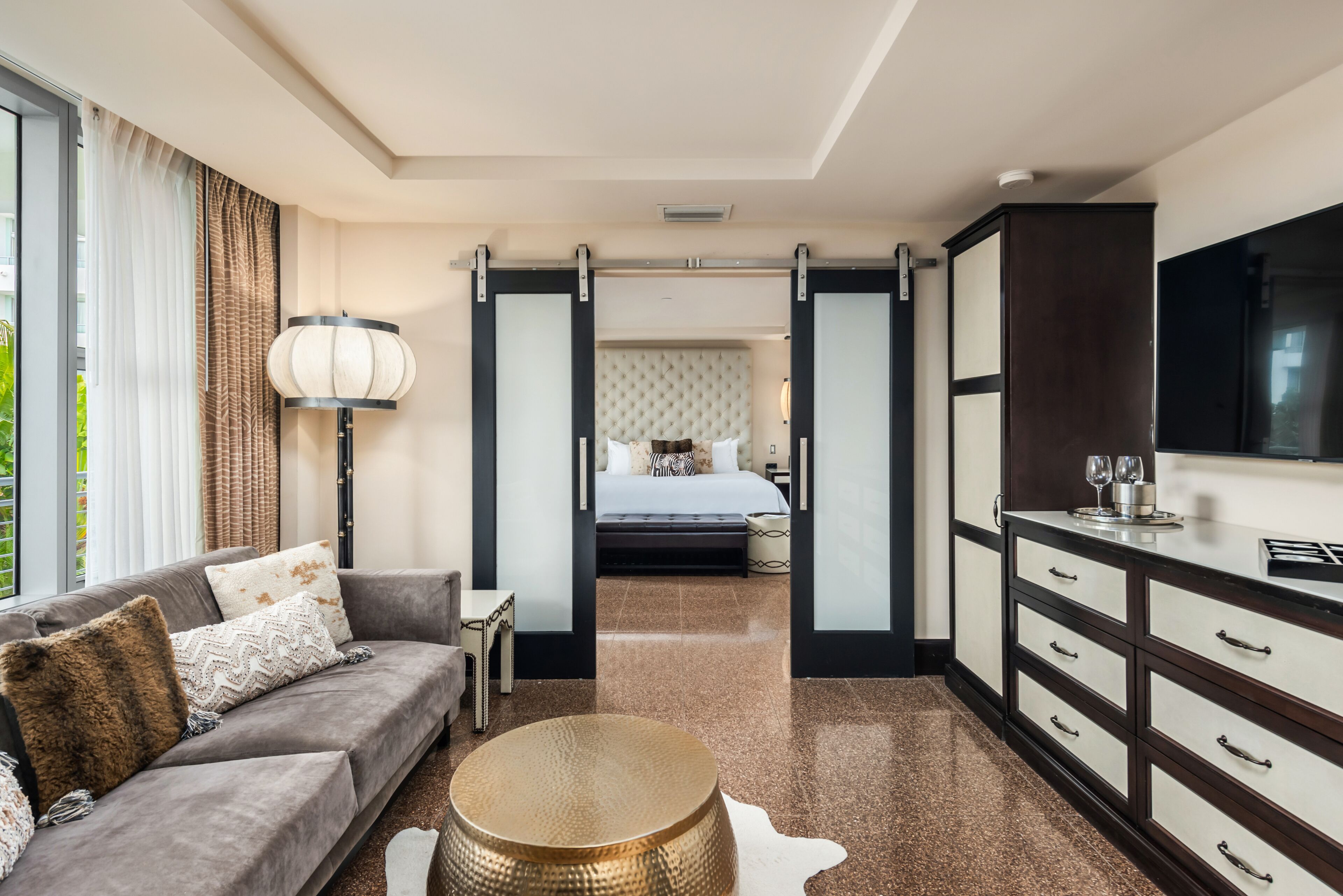 A modern hotel room with a sofa, a round coffee table, a bed with a tufted headboard, a wardrobe, and a large window with curtains.