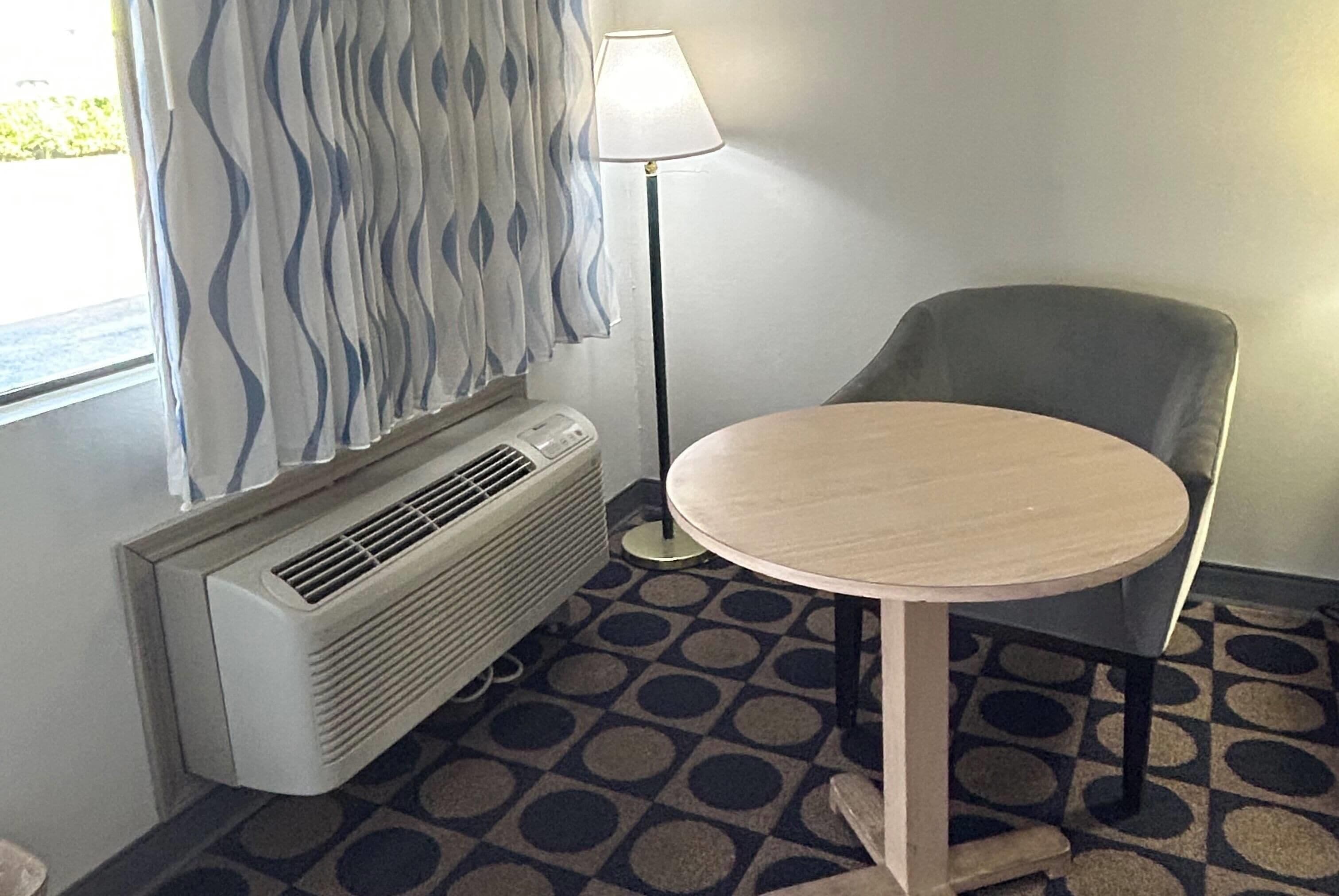 A room with a round table, a chair, a standing lamp, and a window with curtains.