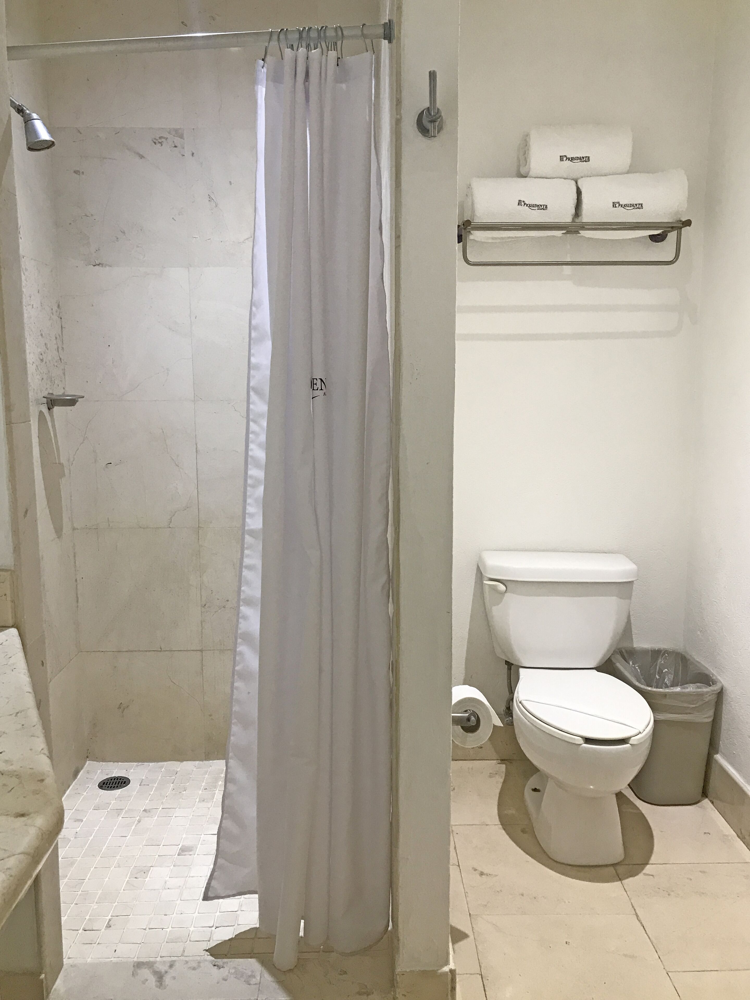 A bathroom with a shower curtain, a toilet, a trash bin, and a shelf with towels.