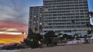 Un hotel de gran altura con varios pisos, una playa en primer plano y palmeras.