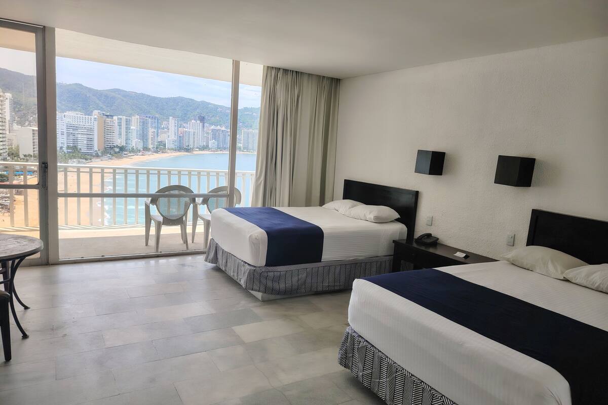 A hotel room with two beds, a balcony with a view of the beach and cityscape, a small table, and a chair.