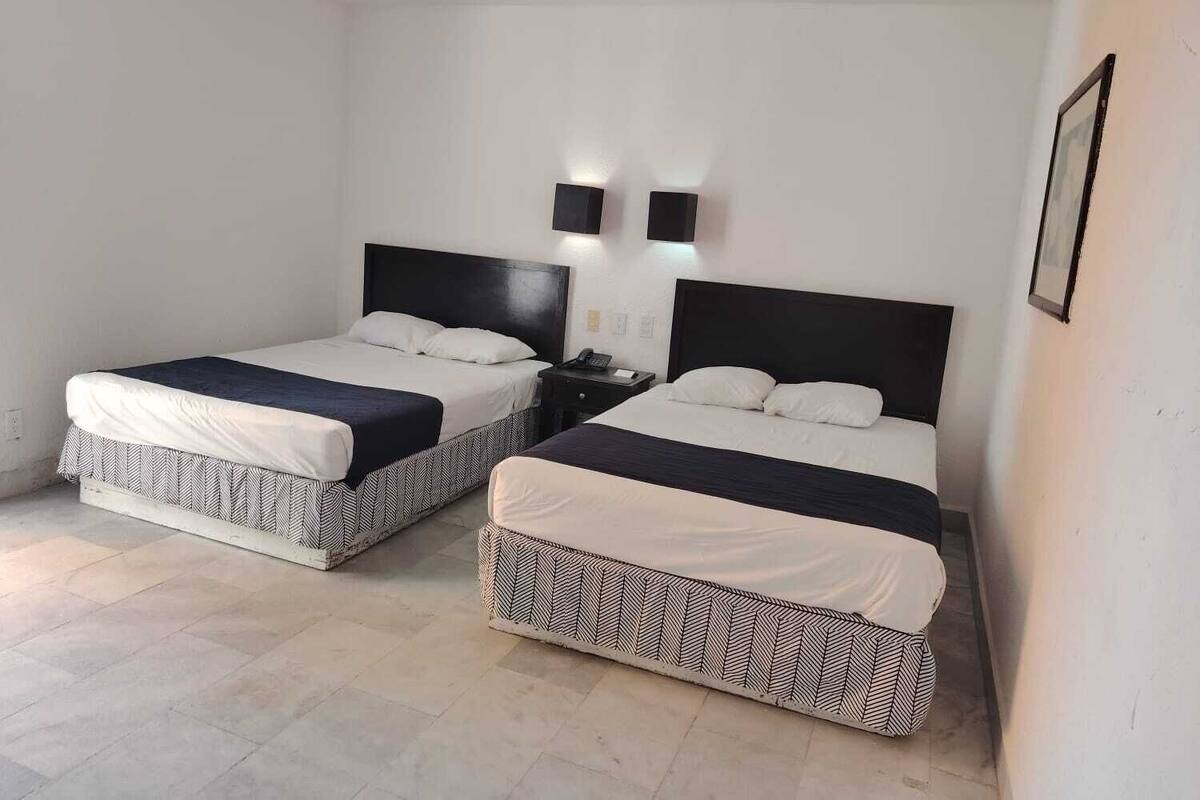 A hotel room with two beds, each with a black headboard and white bedding. There are two bedside tables with lamps, and a framed picture on the wall.