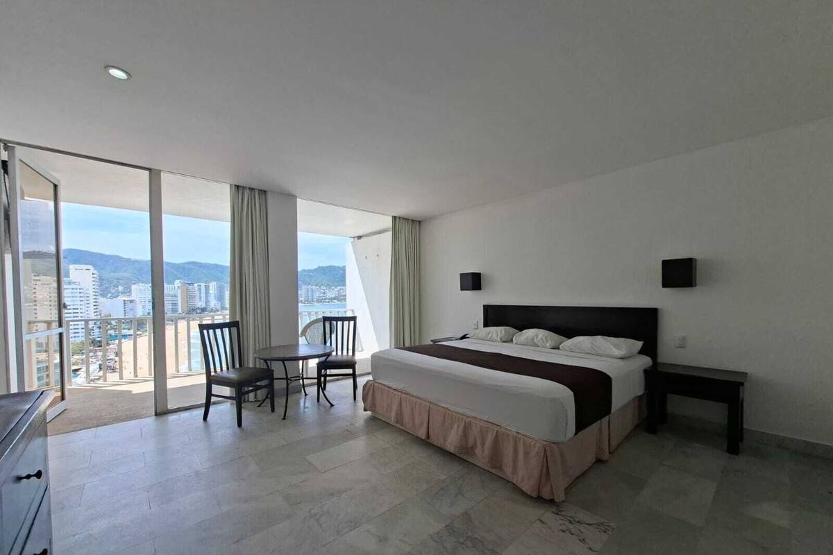 A modern hotel room with a large bed, a dining table, and a view of the city through large windows.