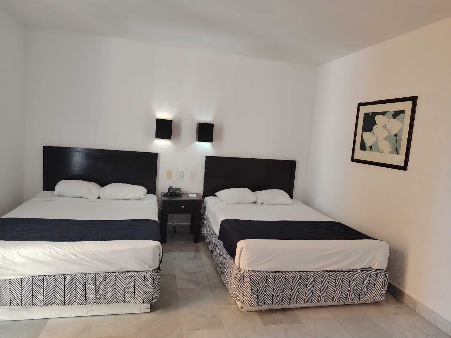 Habitación de hotel con dos camas, una mesita de noche y una lámpara fijada a la pared.