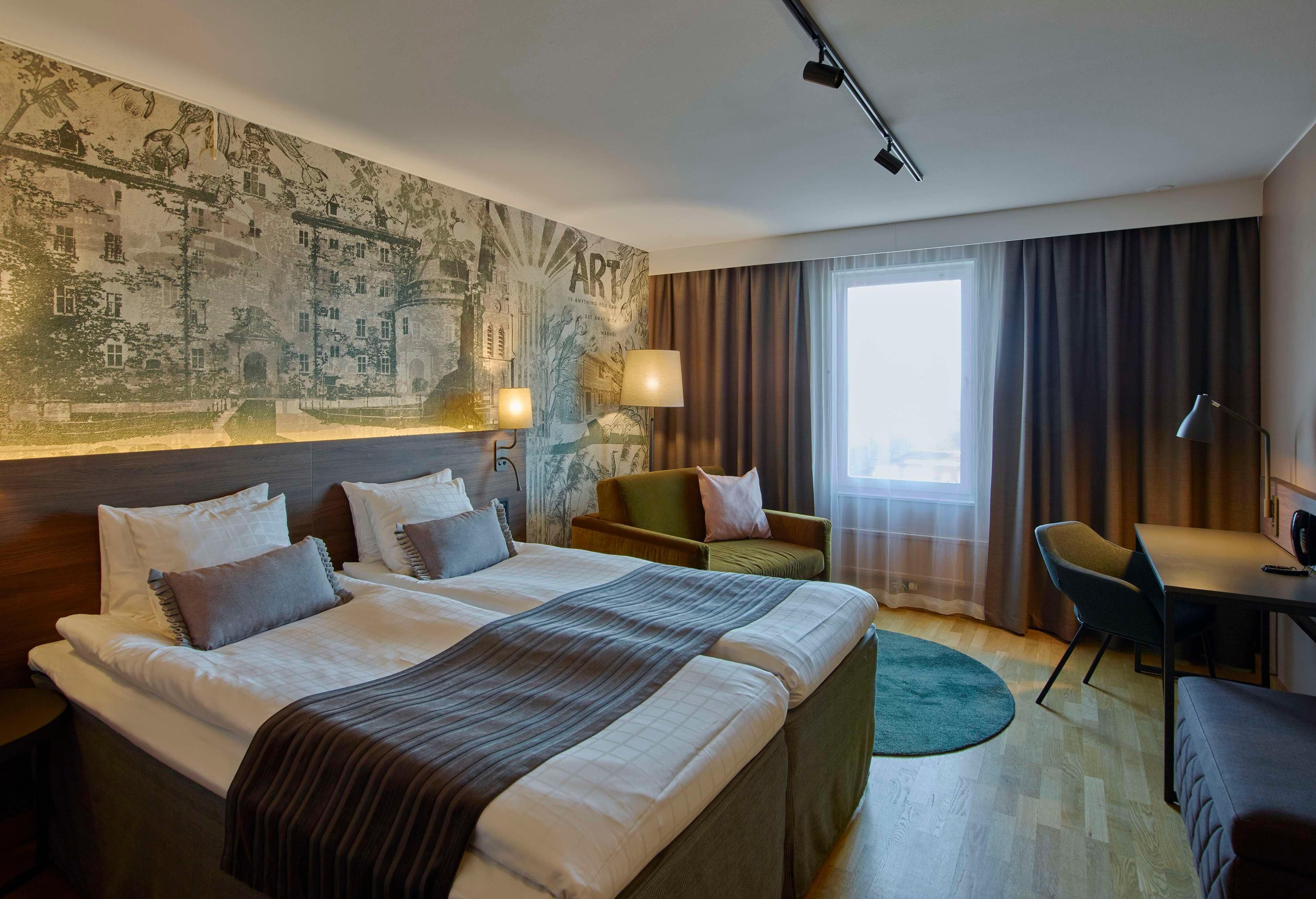Habitación de hotel con dos camas, un escritorio, una silla, un ventanal con cortinas y un mural en la pared.