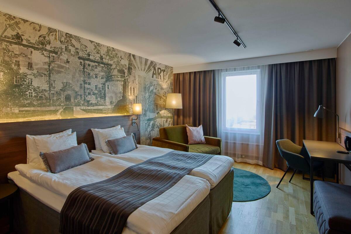 A hotel room with two beds, a desk, a chair, a large window with curtains, and a wall mural.