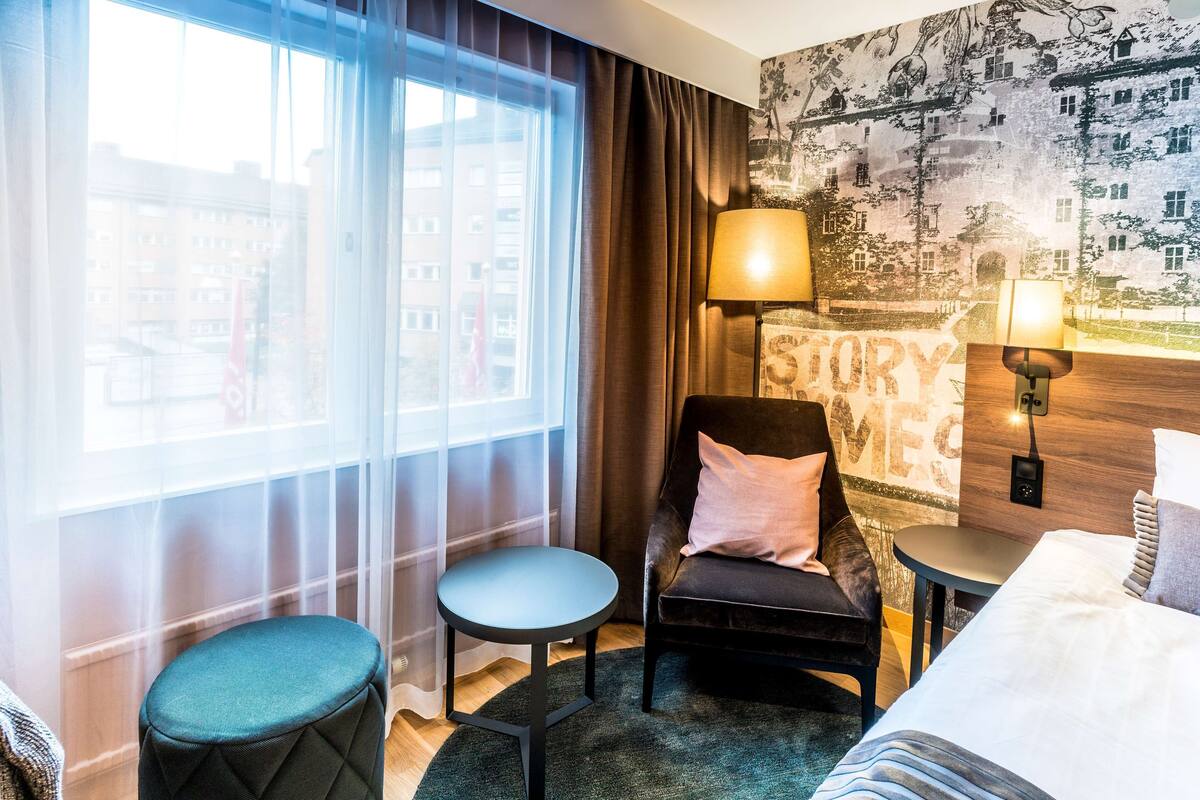 A hotel room with a large window, a chair, a small table, a bed, and a wall with a 'STORY' mural.