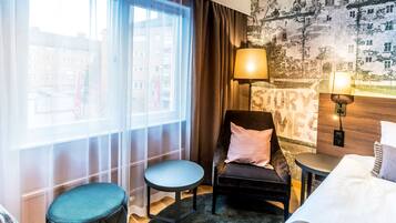 A hotel room with a large window, a chair, a small table, a bed, and a wall with a 'STORY' mural.