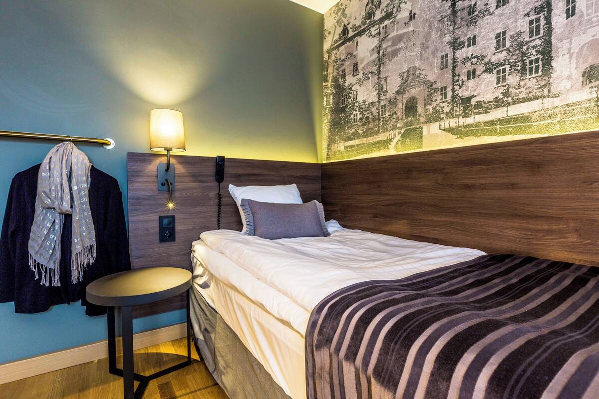 A hotel room with a bed, a nightstand, a lamp, a wall-mounted phone, and a towel rack.